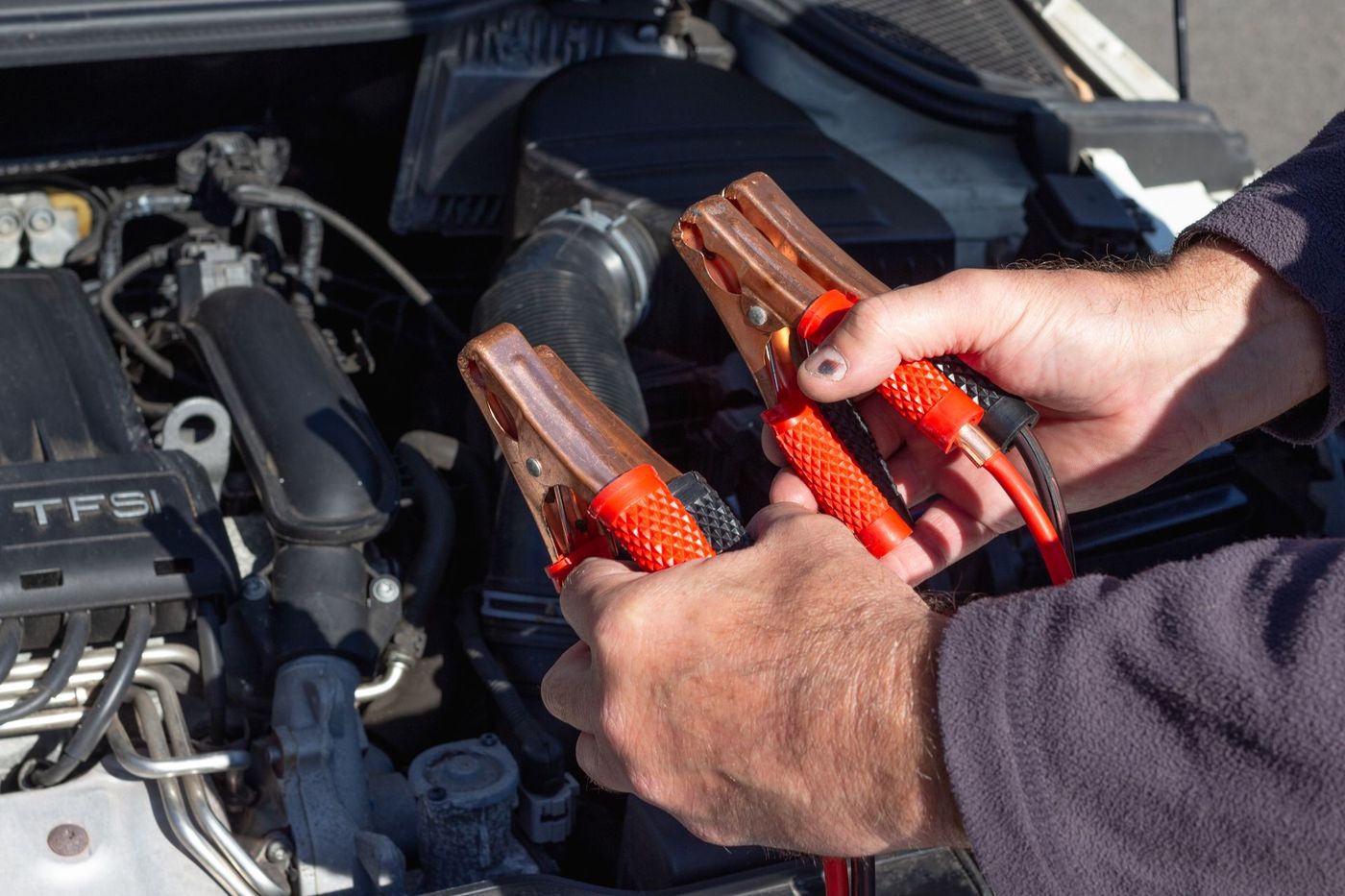 Hands holding jumper cable clamps over an open engine bay outdoors