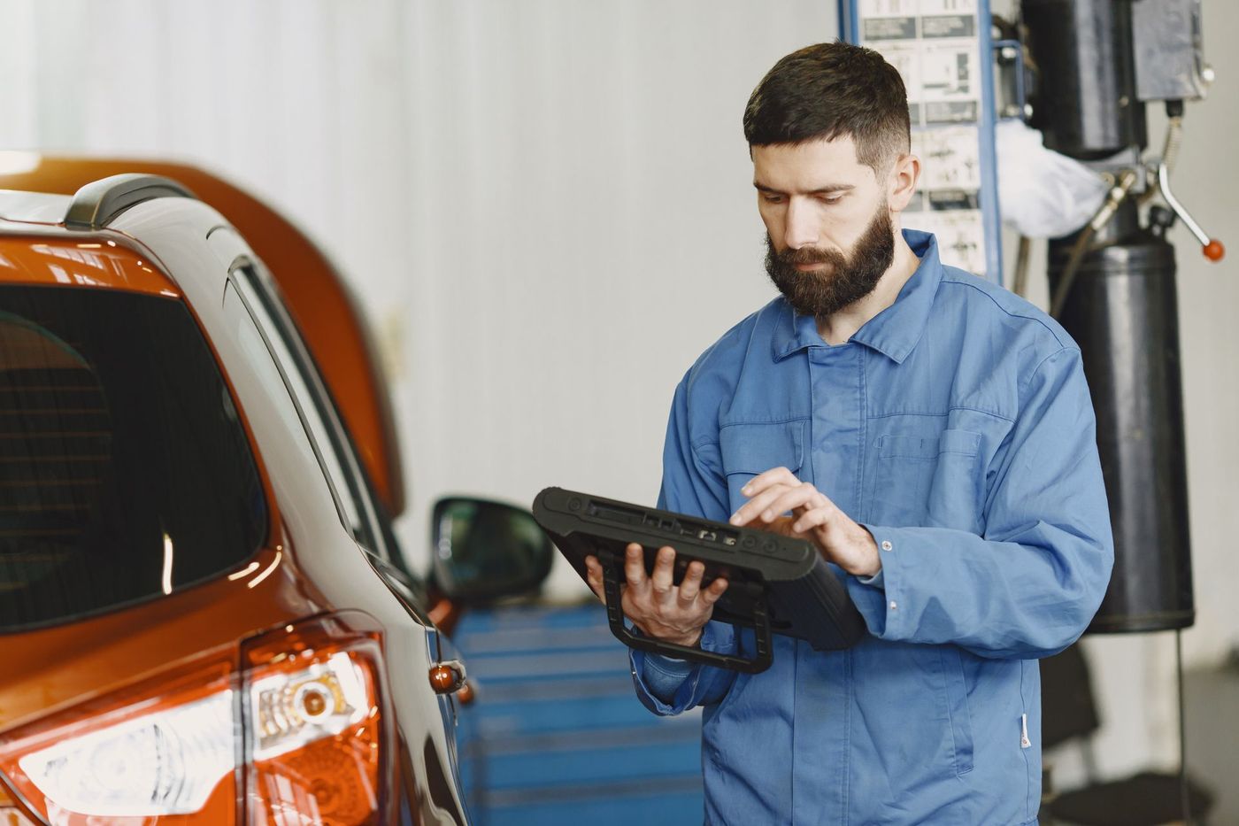 Technician in plain coveralls reading a handheld automotive diagnostic tool beside a car