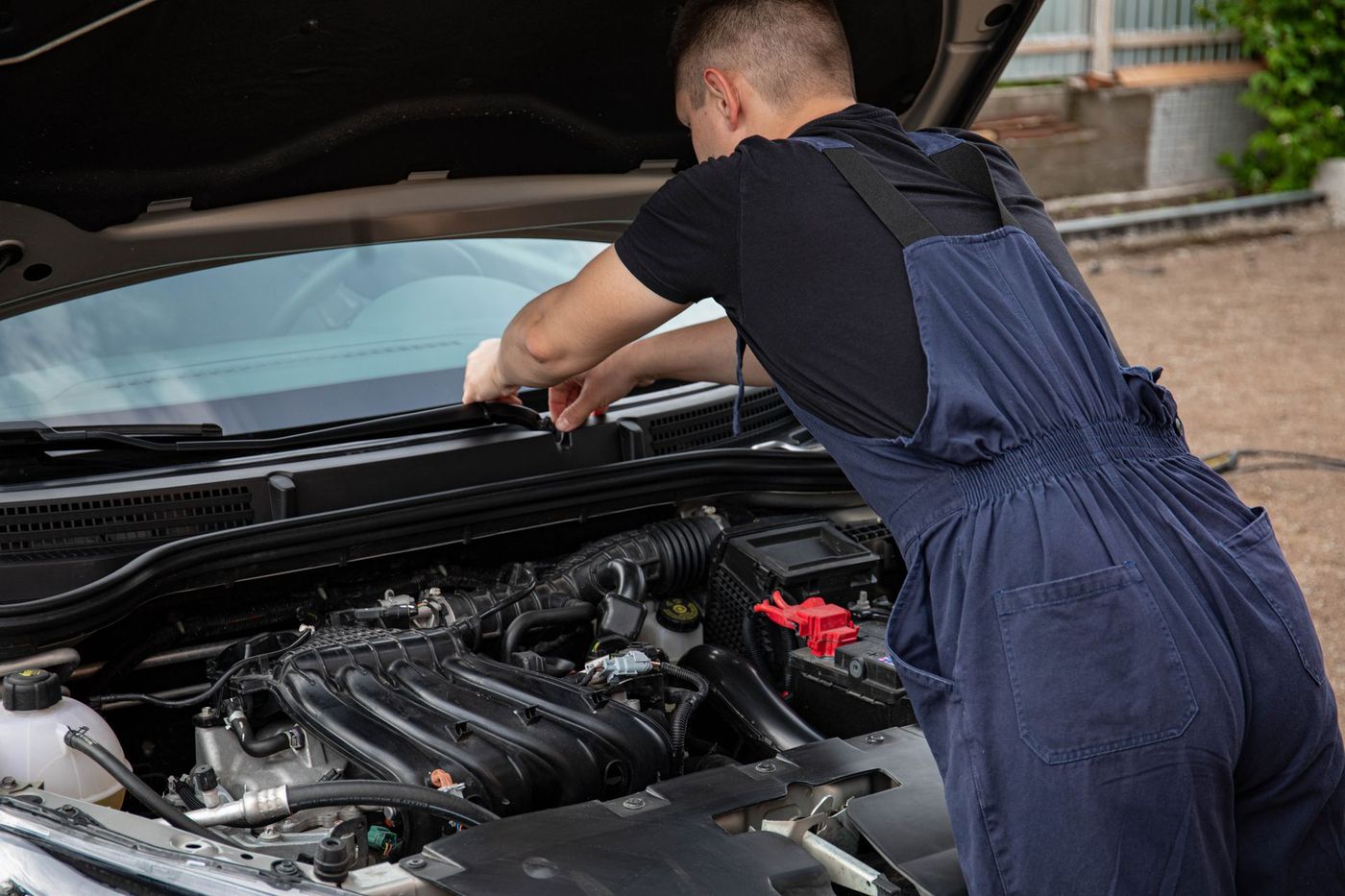 Mechanic in overalls leaning into an open hood, working outdoors on gravel